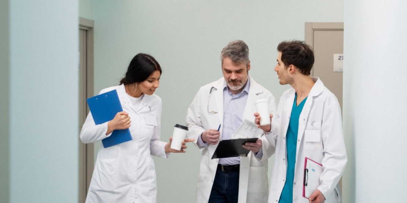 Garde médecin : assurer la continuité des soins Trois médecins échangent dans le couloir d'un établissement de santé lors d'une garde médicale, illustrant la coordination des équipes et la continuité des soins.
