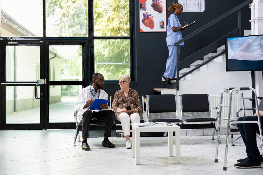 Médecin discutant avec une patiente âgée dans la salle d'attente d'une clinique pendant qu'une infirmière travaille à proximité.